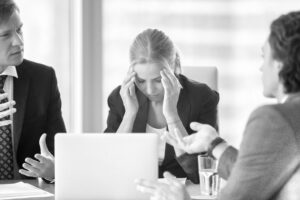 A black and white photo of a business woman sitting in a meeting with her laptop looking down and holding her head as if she has a headache.