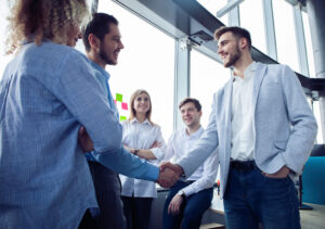 A color photo of several people gathered around two businessmen shaking hands.