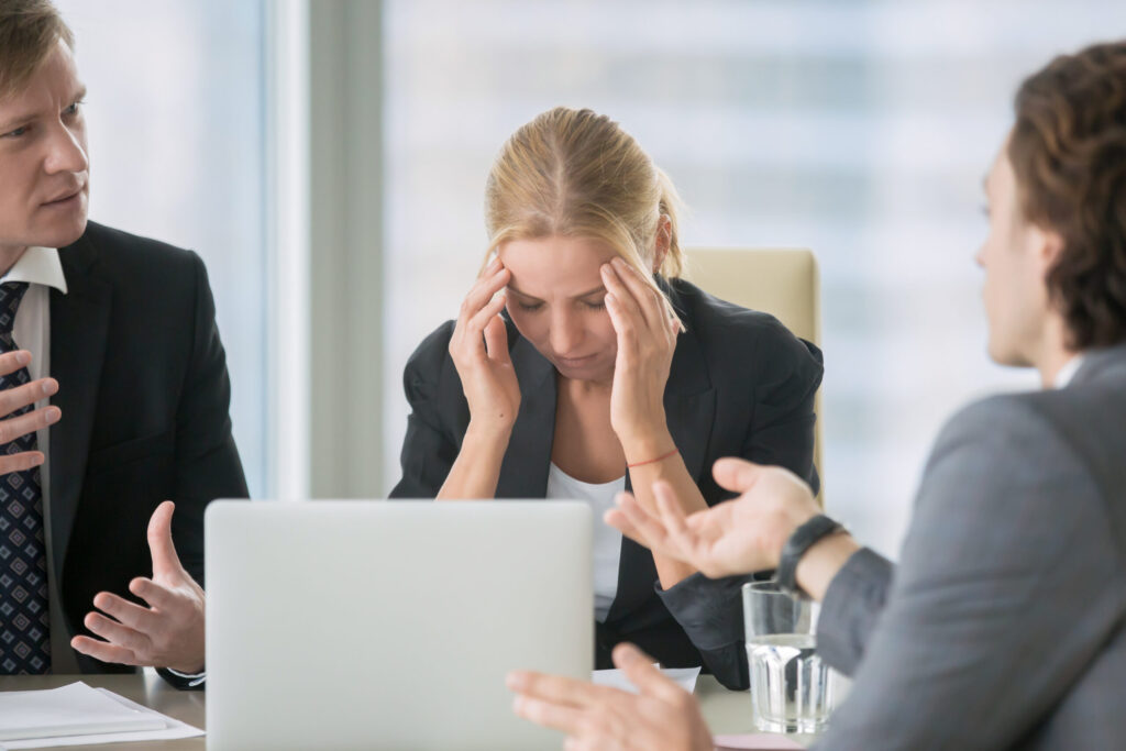 A color photo of a business woman sitting in a meeting with her laptop looking down and holding her head as if she has a headache.